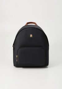 Sac à dos noir avec une finition en tissu lisse, forme arrondie, poignée supérieure marron, poche avant zippée et détail de logo doré à l'avant.