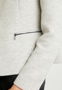 Veste gris clair texturée avec un subtil motif chevrons, dotée d'une poche zippée latérale avec des garnitures argentées et d'une coupe ajustée.