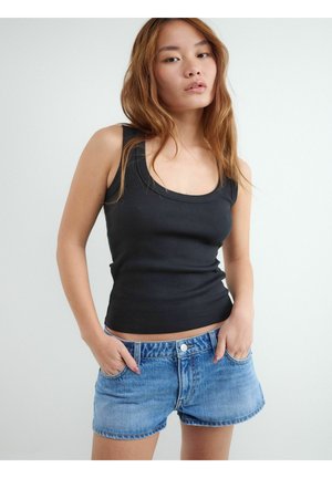 Młoda kobieta ubrana w czarny top bez rękawów i niebieskie krótkie dżinsy, stoi z rękami w kieszeniach na jasnym, jednolitym tle.