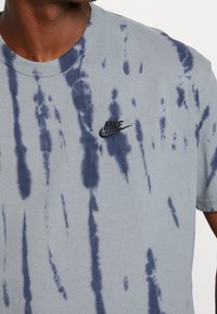 Šedé tričko s tie-dye vzorem a námořnickými modrými skvrnami, kulatý výstřih, krátké rukávy a malá černá vyšitá značka Nike na hrudi.