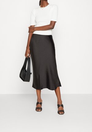 Nainen, jolla on valkoinen lyhythihainen paita, musta satiininen midihame, mustat korolliset sandaalit ja musta käsilaukku valkoista taustaa vasten.