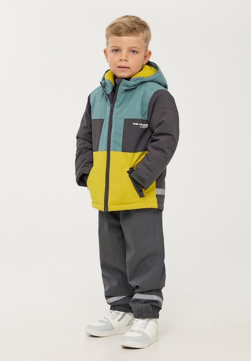 Giacca a blocchi di colore in teal, grigio e giallo con cappuccio; materiale isolante; abbinata a pantaloni grigi; sneakers bianche completano il look.