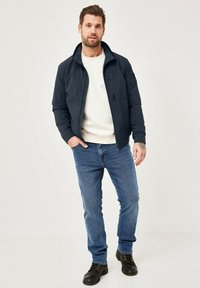 Navy jacket with front zip and pockets, paired with a cream sweater and blue jeans. Black boots complete the outfit. Smooth, casual design.