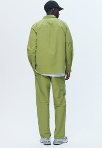 Green long-sleeve button-up shirt and matching trousers in light fabric, with a relaxed fit. Worn with a black cap and gray sneakers.