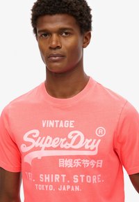 T-shirt en coton corail avec un col ras du cou ; présente un texte blanc affichant "Vintage SuperDry" et des caractères japonais. Manches courtes, coupe décontractée.