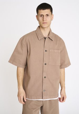 Ung mand iført en løs, kortærmet beige skjorte med knapper og brystlomme, stående mod en ensfarvet hvid baggrund.