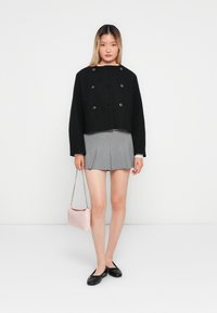Femme se tenant devant un fond blanc, portant une veste noire à double boutonnage courte, une jupe plissée grise, des ballerines noires, et tenant un sac à main rose.