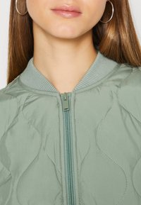Miętowa kurtka typu bomber z pikowanym wzorem, prążkowanym kołnierzem i zapięciem na zamek błyskawiczny. Gładka tekstura z kontrastującymi detalami szwów.