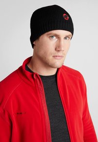 Black ribbed beanie with red logo. Model wears a red fleece jacket over a black shirt. Focus on texture and color contrast.