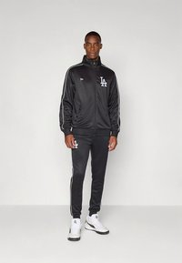 Črna atletska trenirka iz gladkega blaga. Ima bele črte ob straneh in logotip na prsih in nogi. Oblečena z belimi supergami.