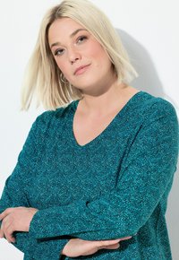 Teall V-ringad topp med långa ärmar, med en strukturerad mönster av små prickar. Tyget verkar mjukt och något elastiskt.