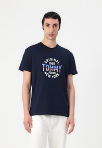 Navy blue cotton t-shirt featuring "Tommy Jeans New York" graphic in red, white, and blue. Short sleeves and crew neckline.