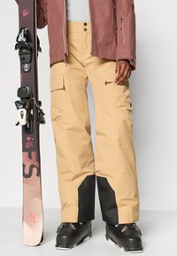 Calças de esqui bege com bolsos cargo, detalhes pretos nas bainhas, combinadas com botas de esqui pretas. Prancha de snowboard segurada com uma mão.