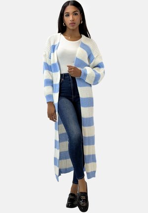 Lange cardigan met verticale blauwe en witte strepen; chunky brei textuur; open ontwerp; gecombineerd met strakke jeans en loafers.