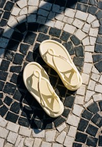 Paire de sandales beiges sur un pavé en pierre à motif noir et blanc avec une ombre visible en haut à gauche.