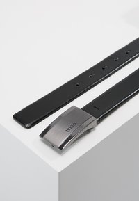 Ceinture en cuir noir avec une boucle en métal chromé portant un logo gravé. La sangle dispose de plusieurs trous de taille pour un ajustement.