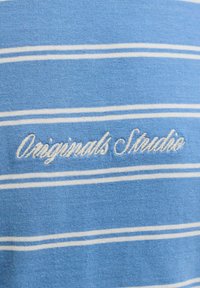 Tissu bleu clair avec de fines rayures blanches horizontales et texte brodé en blanc indiquant "Originals Studio".