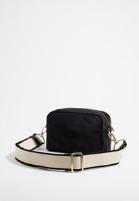 Sort stof crossover tas med rektangulær form, guld lynlås og en bred beige rem med sort kant.