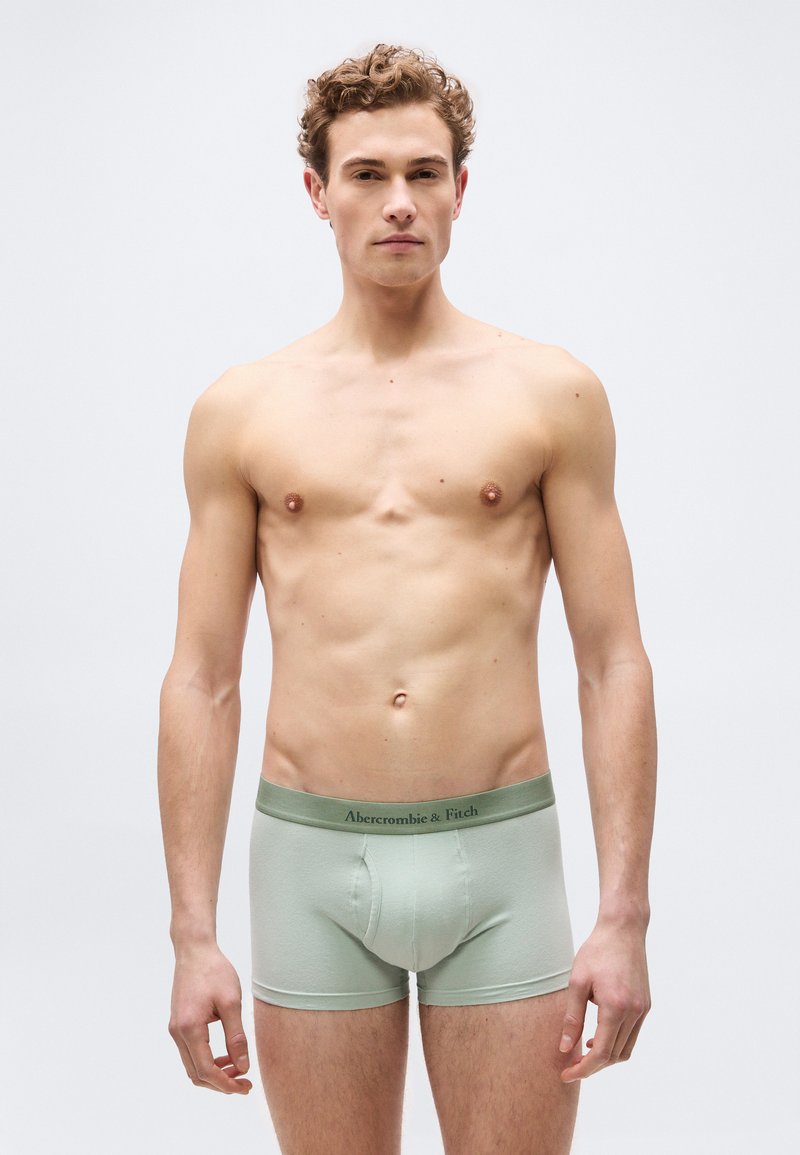 Νεαρός άνδρας με σγουρά μαλλιά, όρθιος, φορώντας ανοιχτοπράσινα εσώρουχα boxer Abercrombie & Fitch, μπροστά σε απλό ανοιχτόχρωμο φόντο.