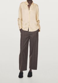 Chemise en satin jaune clair à boutons, associée à un pantalon large marron et à des chaussures à enfiler noires texturées. La chemise présente un col et une coupe décontractée.