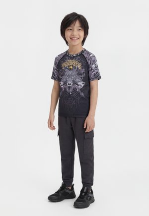 Garçon souriant portant un T-shirt graphique noir avec un motif de loup à trois têtes, un pantalon cargo foncé et des baskets noires, debout sur un fond blanc.