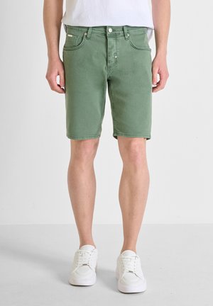Pantalones cortos de mezclilla verde con corte recto, que cuentan con bolsillos frontales y un cierre de botón, combinados con zapatillas blancas sobre un fondo neutral.