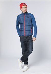 Blaue gesteppte Jacke mit roten Reißverschlüssen und Seitentaschen, kombiniert mit dunkler Jeans und weißen Turnschuhen. Das Modell trägt eine rote Mütze.