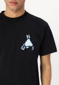 Black cotton t-shirt featuring a white and blue graphic design with the letters "H," "U," and "F" and a triangular shape on the left chest.