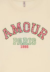 Kreminė marškiniai su užrašu "AMOUR" raudonais leopardų raštu, "PARIS" žaliais blokiniais raidėmis ir "1995" raudonai apačioje.