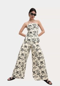 Strapless top en wijde broekset in crÃ¨me met een zwart bloemenpatroon. Heeft een gladde textuur en een subtiele glans. Zwarte sandalen maken de look af.
