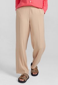 Beige vida byxor med slät yta, med framveck och skräddarsydd passform. Matchas med bruna slip-in sandaler.