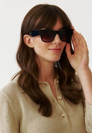 Okulary przeciwsłoneczne z czarną oprawką, ciemnymi gradientowymi szkłami, kwadratowym kształtem i subtelnymi bocznymi zdobieniami. Noszone przez osobę z długimi włosami w beżowym swetrze.