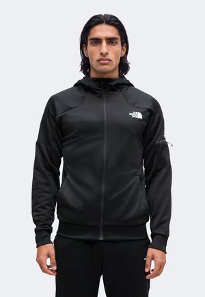 Hombre con pelo negro lleva una sudadera negra con cremallera de The North Face con el logo en el pecho, de pie mirando hacia adelante frente a un fondo liso y claro.