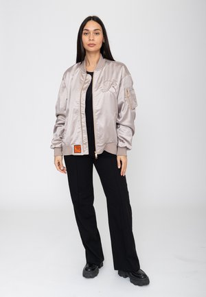 Frau steht und trägt eine hellgraue Satin-Bomberjacke, schwarze Hose und schwarze klobige Schuhe vor einem schlichten weißen Hintergrund.