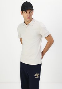 Helles beiges Polohemd mit weichem Textil, ausgestattet mit einem Logo auf der Brust und am Ärmel. Kombiniert mit marineblauen Jogginghosen mit goldenen Akzenten.