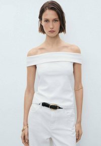 Witte off-the-shoulder top van gladde stof, gecombineerd met witte broek en een zwarte riem met een gouden accent. Eenvoudig ontwerp, geen patronen.