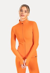 Orangene Zip-Jacke für Sport mit langen Ärmeln und Daumenlöchern, aus dehnbarem Material gefertigt. Flauschige Nähte und ein hoher Kragen.