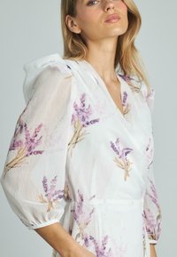 Weißes Kleid mit lavendelfarbenem Blumendruck, das puffärmel und einen V-Ausschnitt hat. Aus leichtem, strukturiertem Stoff gefertigt.