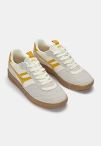Weiße und gelbe Sneakers aus Wildleder und Leder, mit perforiertem Zehenbereich, flachen Schnürsenkeln und einer Gummisohle. Gelbe Akzente an den Seiten.