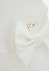 Weißes Kleid mit großer Satinschleife an der Taille, akzentuiert mit einem goldenen "Guess"-Logo. Das Material wirkt weich und hat eine Tüll-Überlagerung.
