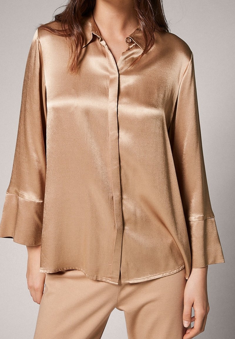 Blusa in satin beige con colletto, chiusura con bottoni e maniche a campana 3/4. Tessuto liscio con una leggera lucentezza e un taglio morbido.