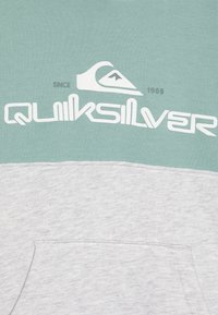 Svetlozelena i melirana siva dukserica s kengurufrontom, s istaknutim bijelim logotipom "Quiksilver" i detaljima dizajna iz 1969. godine.