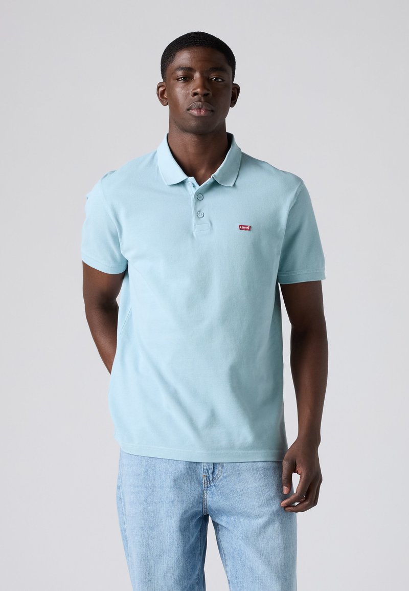 Homme debout, les mains dans le dos, portant un polo Levi's bleu clair et un jean bleu clair sur un fond gris.