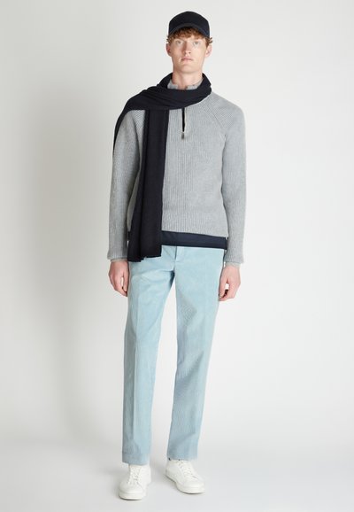 Grijze gebreide trui met een hoge kraag en rits, gecombineerd met lichtblauwe gestreepte broek en witte sneakers. Zwarte sjaal over de schouders gedrapeerd.