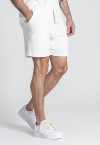 Pantalones cortos blancos con textura y bolsillos laterales, combinados con zapatillas blancas con cordones crema. Enfocados en la comodidad y un diseño casual.