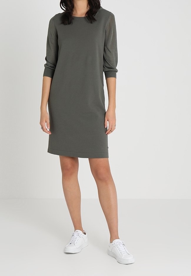 Femme portant une robe kaki longueur genoux avec des manches trois-quarts et des baskets blanches, debout devant un fond clair uni.