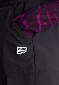 Čierna látková kapsa s bielym vyšívaným logom PUMA. Fialový vzor s ružovými dizajnmi puma vykúka z vnútra kapsy.
