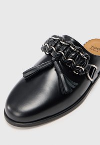 Pantofii negri din piele, cu slip-on, prezintă o bandă împletită cu detalii din lanț metalic și ciucuri. Textură netedă și design cu vârful rotunjit.