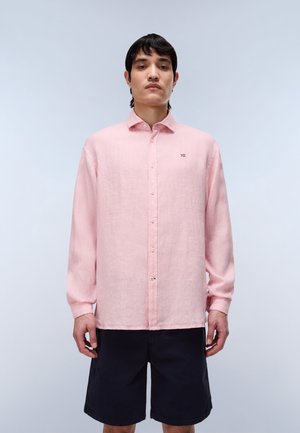 Młody mężczyzna w jasnoróżowej, długorękawowej koszuli z guzikami i czarnych szortach do kolan, stojący na jednolitym tle.