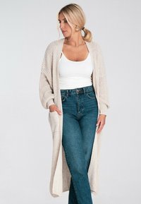 Długi, otwarty kardigan w jasnym beżu z dzianiny, z luźnymi rękawami i kieszeniami, w zestawie z białą koszulką na ramiączkach i dżinsami.